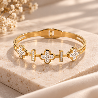 A gold-plated bangle bracelet with three clover motifs and small clear rhinestones, displayed on a beige stone slab with dried flowers in the background.