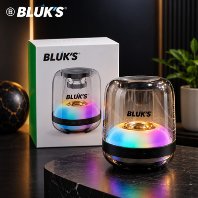 BLUK'S portable Bluetooth speaker with a clear dome-shaped glass body, black base, and multicolor LED light, shown next to its white and green retail box on a dark marble surface.