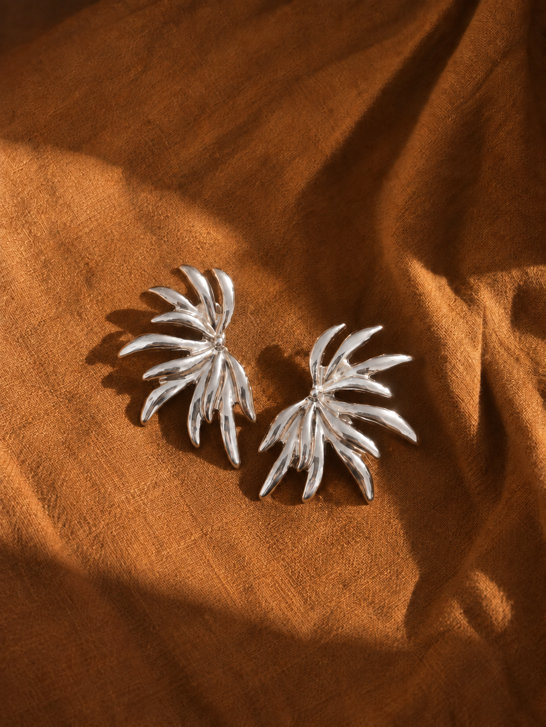 A pair of silver-toned palm leaf stud earrings resting on draped brown fabric.