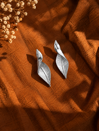 A pair of silver leaf-shaped drop earrings displayed on a textured rust-orange fabric background with dried flowers in the top-left corner.