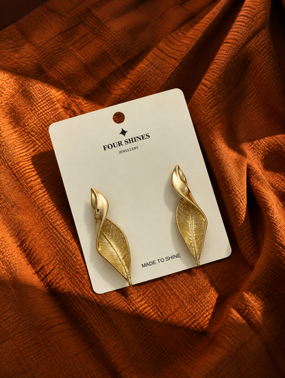 A pair of gold leaf-shaped dangle earrings on a white display card, placed on a textured rust-orange fabric background. The card is branded 'FOUR SHINES JEWELLERY'.