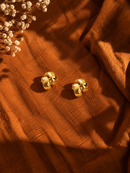 A pair of gold-toned double-huggie hoop earrings resting on a textured rust-orange fabric, styled with dried white flowers in the top-left corner under warm, dramatic sunlight.