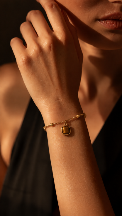 A close-up lifestyle shot of a model's wrist wearing a delicate gold chain bracelet with a small, polished square gold charm.