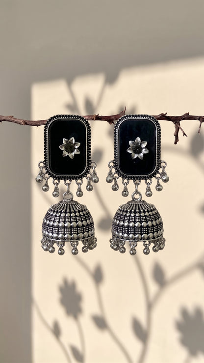 A pair of black and silver jhumka earrings hanging from a thin branch. Each earring features a rectangular black face with a silver floral motif, adorned with dangling silver beads and bell-shaped jhumka drops with intricate silver patterns.