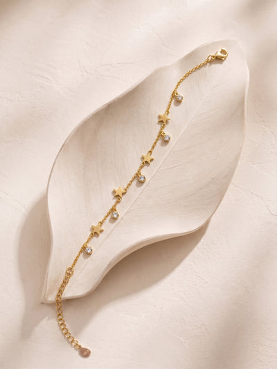 A delicate gold chain bracelet with star and pearl-like bead accents, displayed on a large cream-colored leaf prop against a textured beige background.
