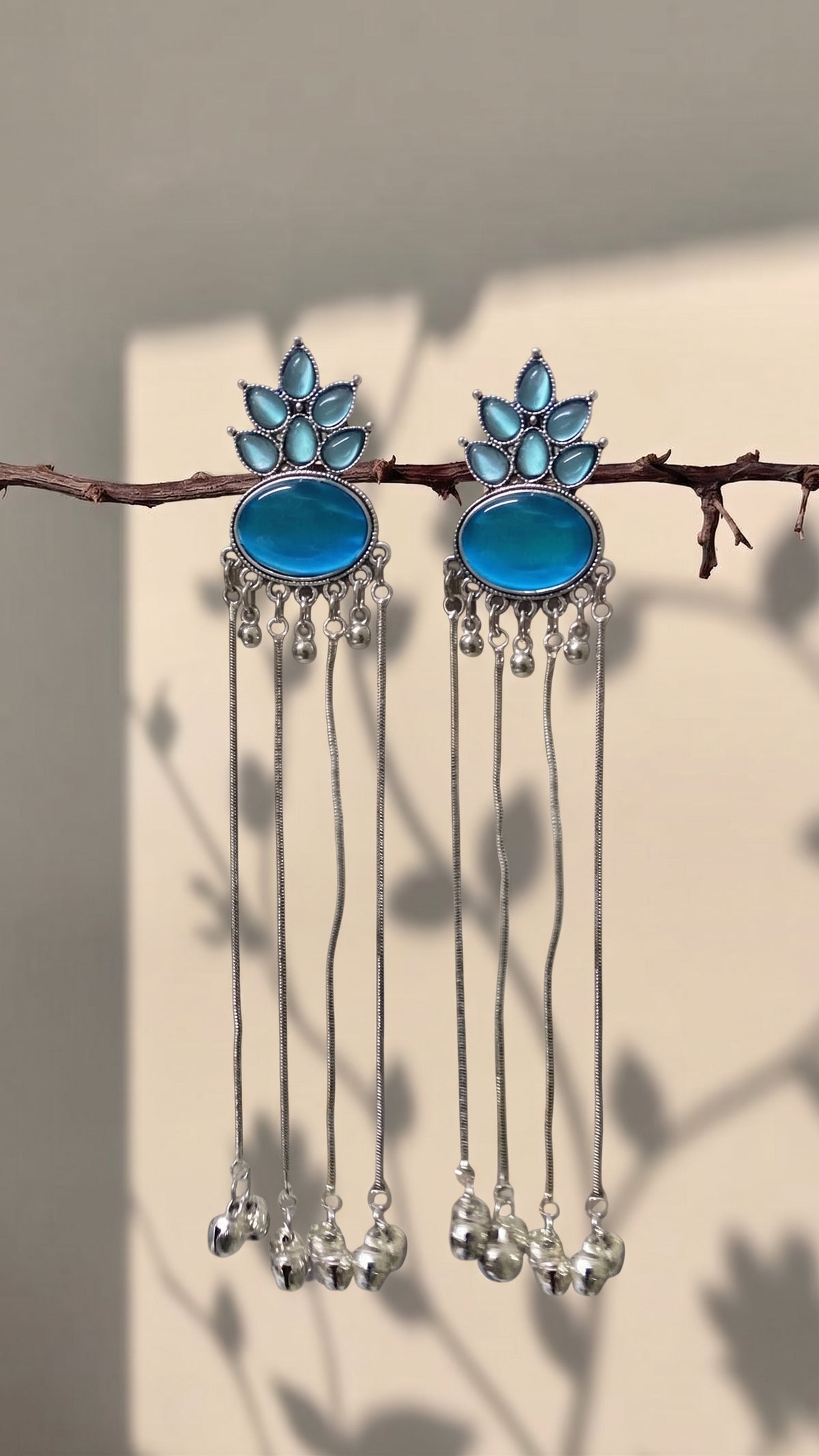 A pair of silver-toned dangle earrings with oval blue stones and leaf-shaped blue accents, hanging from a thin branch against a beige wall with soft leaf shadows.