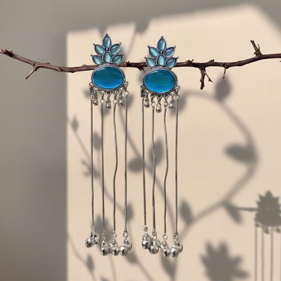 A pair of silver-toned dangle earrings with oval blue stones and leaf-shaped blue accents, hanging from a thin branch against a beige wall with soft leaf shadows.
