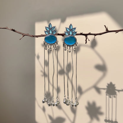 A pair of silver-toned dangle earrings with oval blue stones and leaf-shaped blue accents, hanging from a thin branch against a beige wall with soft leaf shadows.