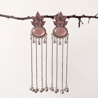 A pair of silver-toned dangle earrings with oval pink stones and small pink accent stones, hanging from a thin branch against a plain off-white background.