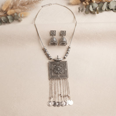 Silver-toned traditional jewelry set featuring a necklace with a square pendant, dangling chains, coin charms, and matching bell-shaped earrings, arranged on a light beige textured surface with dried floral accents in the corners.