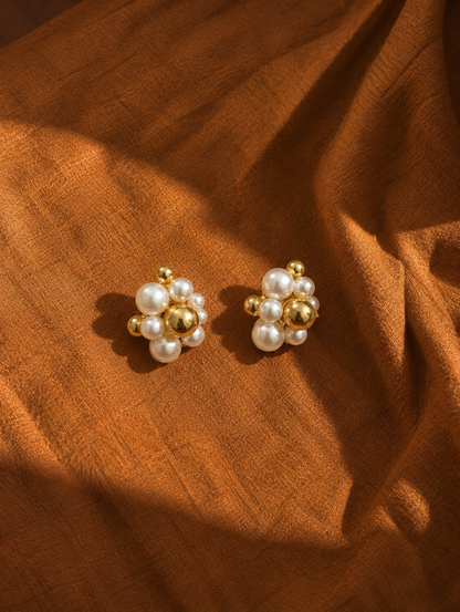 A pair of gold and pearl stud earrings arranged on draped brown fabric.