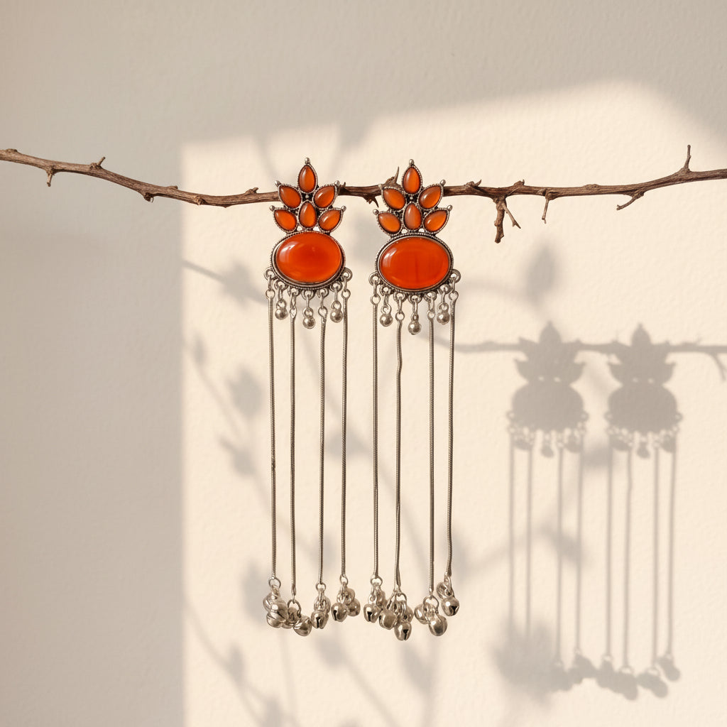 A pair of silver-toned dangle earrings with large orange-red oval stones and smaller orange stones above, hanging from a thin branch against a light wall with soft shadows.