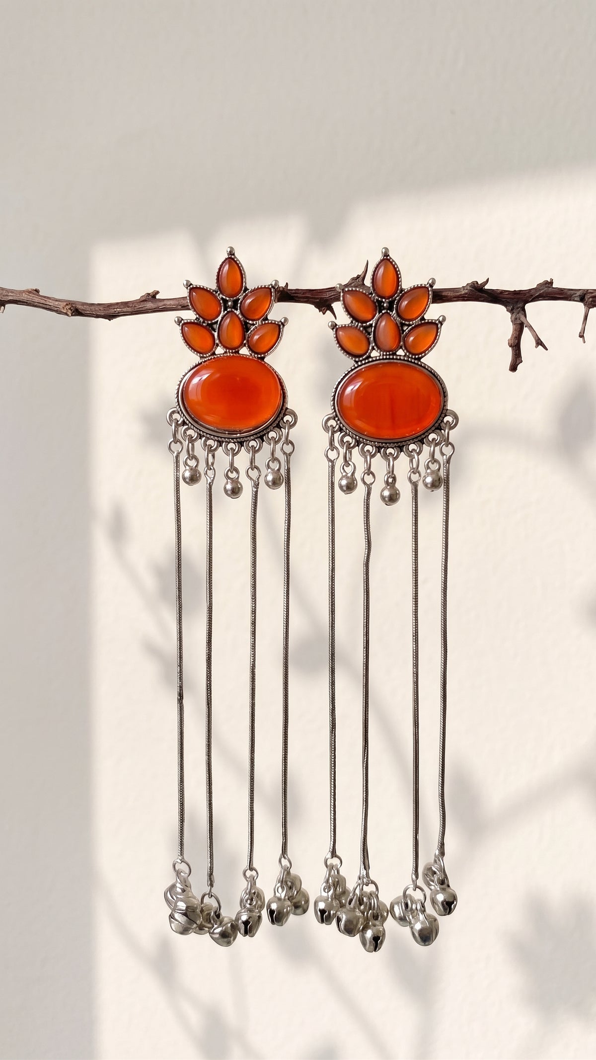 A pair of silver-toned dangle earrings with large orange-red oval stones and smaller orange stones above, hanging from a thin branch against a light wall with soft shadows.