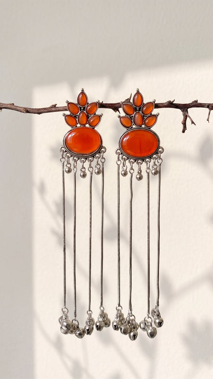 A pair of silver-toned dangle earrings with large orange-red oval stones and smaller orange stones above, hanging from a thin branch against a light wall with soft shadows.