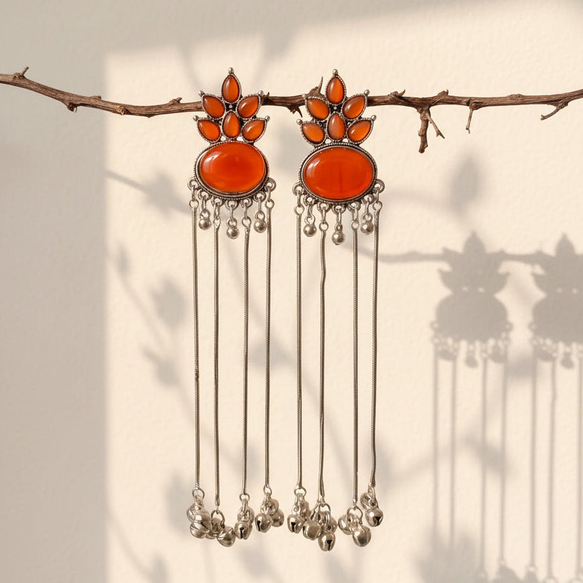 A pair of silver-toned dangle earrings with large orange-red oval stones and smaller orange stones above, hanging from a thin branch against a light wall with soft shadows.