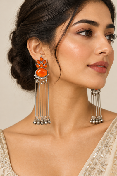 A close-up of a woman wearing ornate silver tassel earrings with bright orange stone accents and dangling silver beads, against a soft beige background.