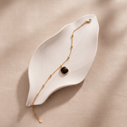 A delicate gold chain bracelet with a central square black cabochon pendant, displayed on a large beige leaf-shaped tray against a textured beige background.