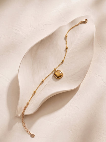 A delicate gold chain bracelet with a small, polished gold square pendant, displayed on a textured, leaf-shaped cream ceramic dish.