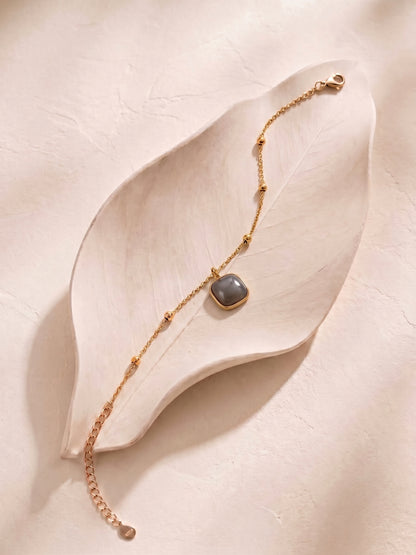 A delicate gold chain bracelet with a square gray stone pendant, displayed on a light beige leaf-shaped tray against a textured beige background.