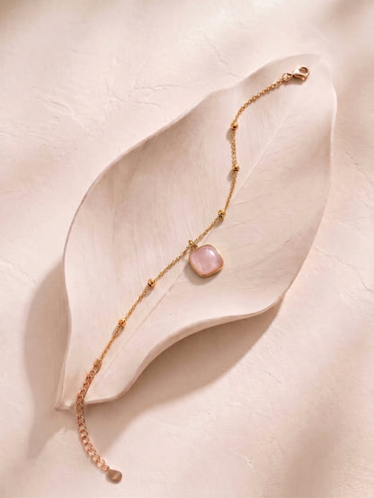 A delicate gold chain bracelet with a small, rounded pale pink stone pendant, styled on a light beige textured leaf-shaped tray.