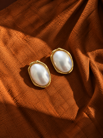 A pair of gold-toned oval earrings with large white pearl-like centers, displayed on draped rust-orange fabric.
