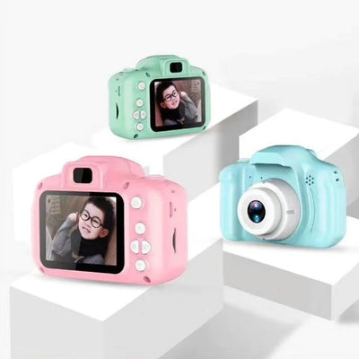 Three children's digital cameras displayed on a stand, with one in pink, one in blue, and one in green. Each camera has a 2.
