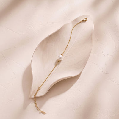 A delicate gold chain bracelet with a single white pearl bead, displayed on a light beige leaf-shaped tray against a textured beige background.