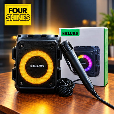 BLUKS portable speaker karaoke system with a built-in microphone, shown on a wooden table next to its packaging box. The speaker features a black rugged casing with a glowing orange ring around the front speaker grille.