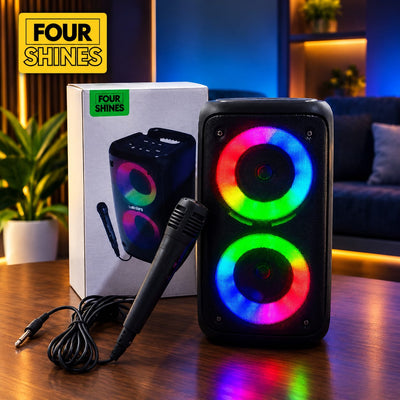 A portable black speaker with two glowing RGB LED rings stands on a wooden table next to its white product box and a black handheld microphone. The background shows a dimly lit living room with blue ambient lighting.