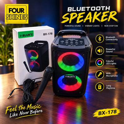 A BLUK'S BX-178 Bluetooth speaker shown on a wooden table with its retail box, microphone, and cables. The speaker features a black body with dual circular LED lights displaying a rainbow spectrum.
