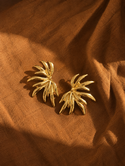 A pair of gold-toned, palm-leaf-shaped earrings resting on draped brown fabric under warm, directional sunlight.