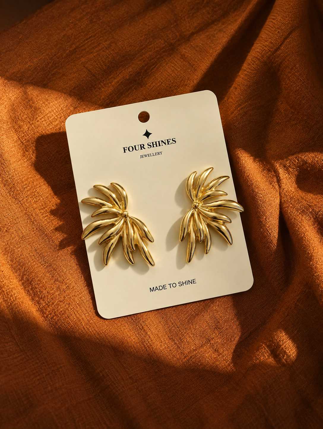 A pair of gold leaf-shaped stud earrings on a white display card labeled 'FOUR SHINES JEWELLERY', placed on a textured rust-orange fabric background.
