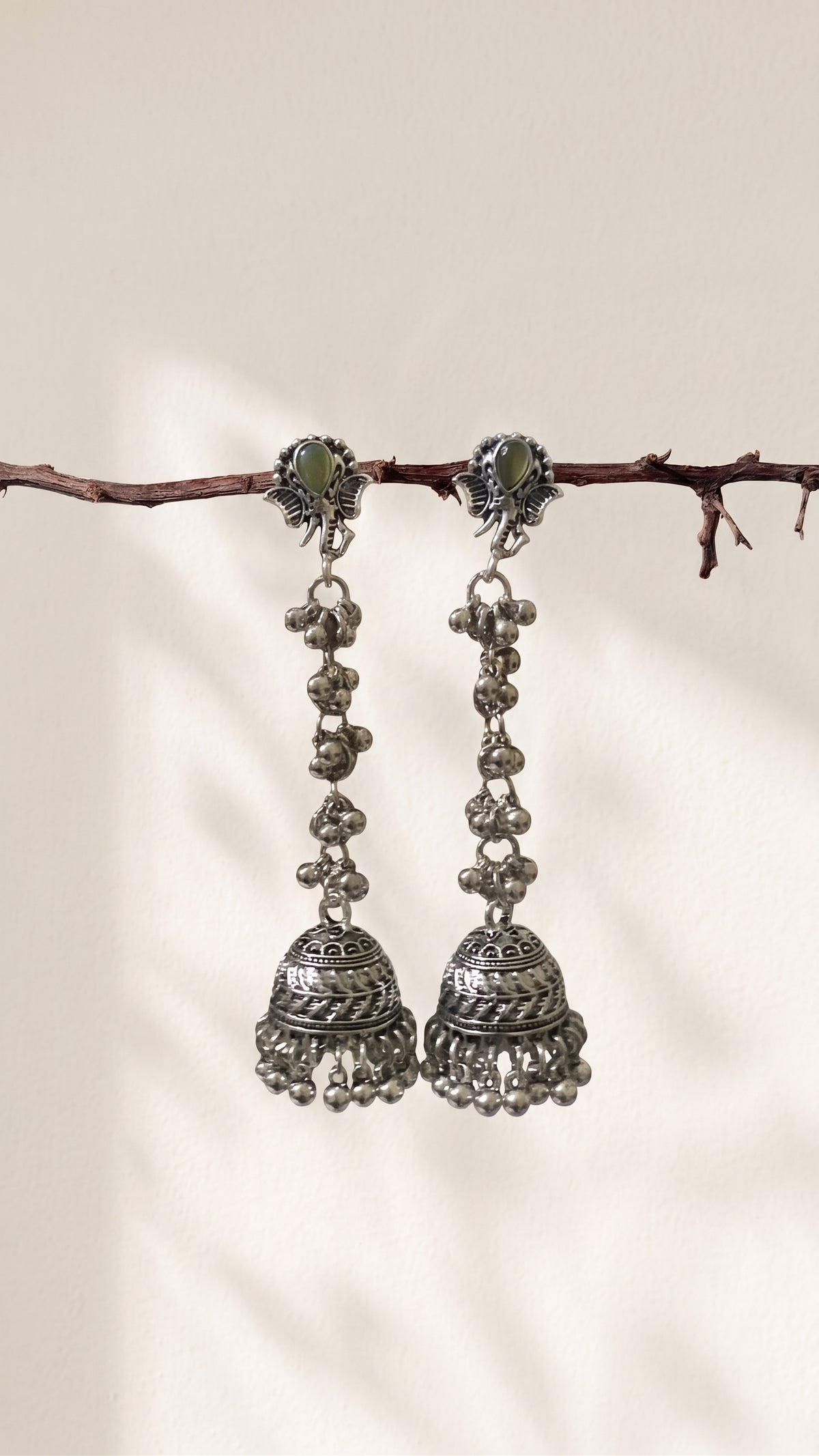 A pair of traditional silver-toned jhumka earrings with green stone accents, hanging from a thin brown branch against a warm off-white background.