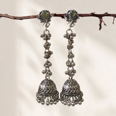 A pair of traditional silver-toned jhumka earrings with green stone accents, hanging from a thin brown branch against a warm off-white background.