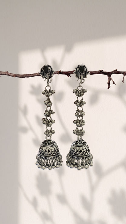 A pair of silver-toned Indian jhumka earrings hanging from a thin brown branch against a warm off-white background with soft leaf shadows.