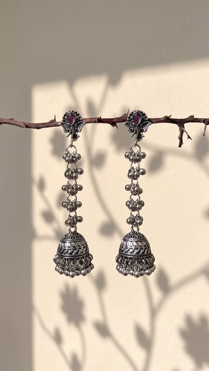 A pair of silver-toned Indian jhumka earrings with small purple stones at the top, hanging from a thin brown branch against a warm beige wall with leaf shadows.