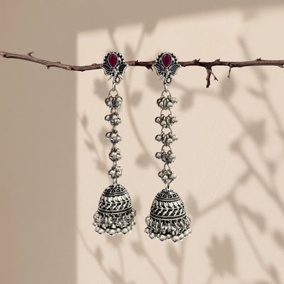 A pair of traditional Indian silver-toned dangle earrings with red oval stones and intricate bell-shaped charms, hanging from a thin branch against a warm beige background with soft leaf shadows.