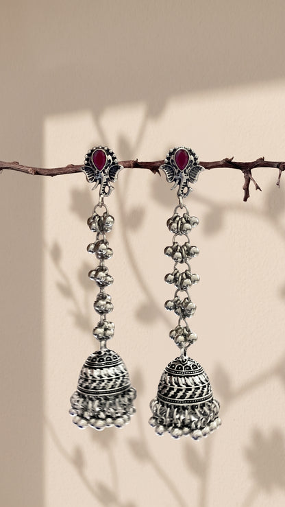 A pair of traditional Indian silver-toned dangle earrings with red oval stones and intricate bell-shaped charms, hanging from a thin branch against a warm beige background with soft leaf shadows.
