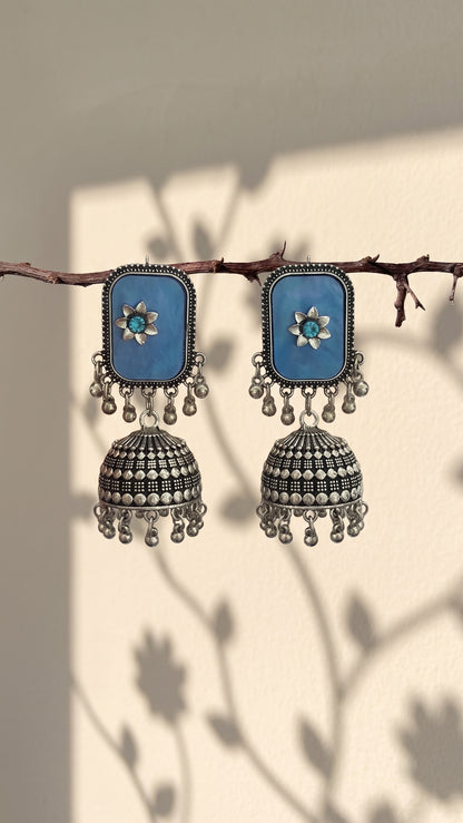 A pair of silver-toned Indian jhumka earrings with rectangular blue stone panels and small turquoise-like blue center stones, hanging from a thin branch against a warm beige wall with leaf shadows.