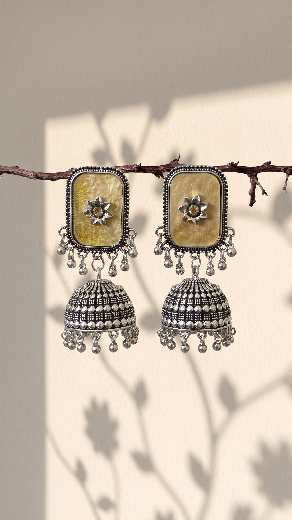 A pair of traditional Indian silver jhumka earrings with rectangular gold-toned mother-of-pearl inlays and dangling silver bells, hanging from a thin branch against a beige wall with leaf shadows.