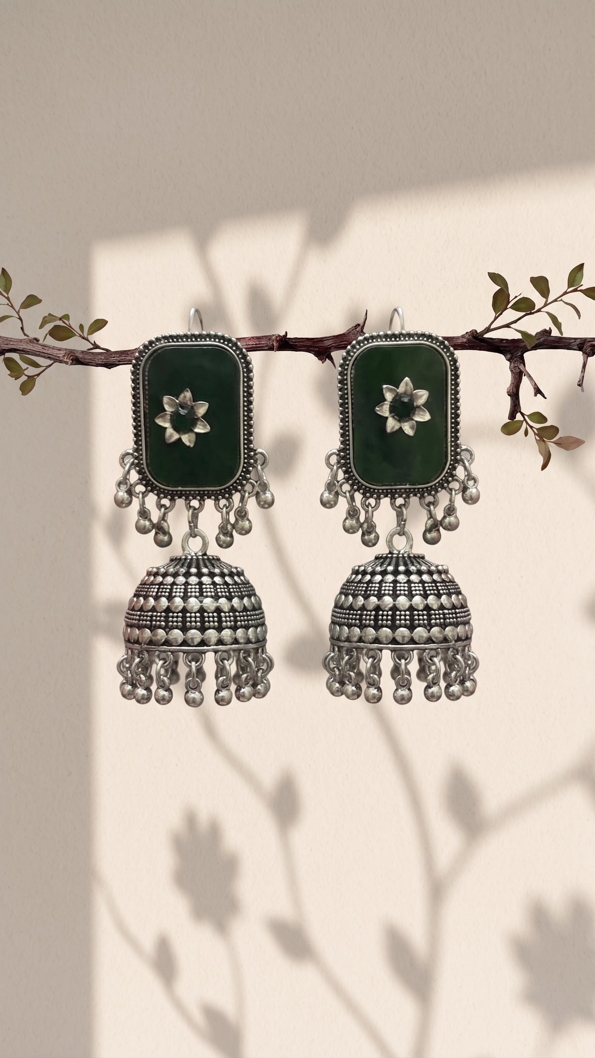 A pair of traditional Indian jhumka earrings hanging from a decorative branch. The earrings feature dark green rectangular inlays with white floral motifs, surrounded by silver-toned borders and dangling silver-toned bell-shaped charms with bead fringe.