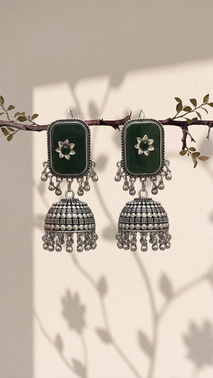 A pair of traditional Indian jhumka earrings hanging from a decorative branch. The earrings feature dark green rectangular inlays with white floral motifs, surrounded by silver-toned borders and dangling silver-toned bell-shaped charms with bead fringe.