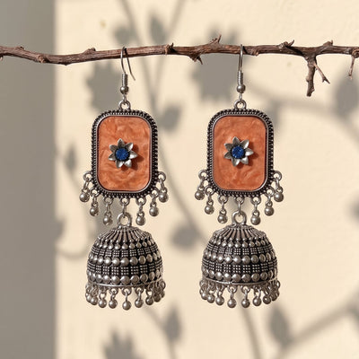 A pair of bohemian-style silver jhumka earrings with rectangular orange stone inlays and blue central stones, hanging from a rustic branch against a wall with leaf shadows.