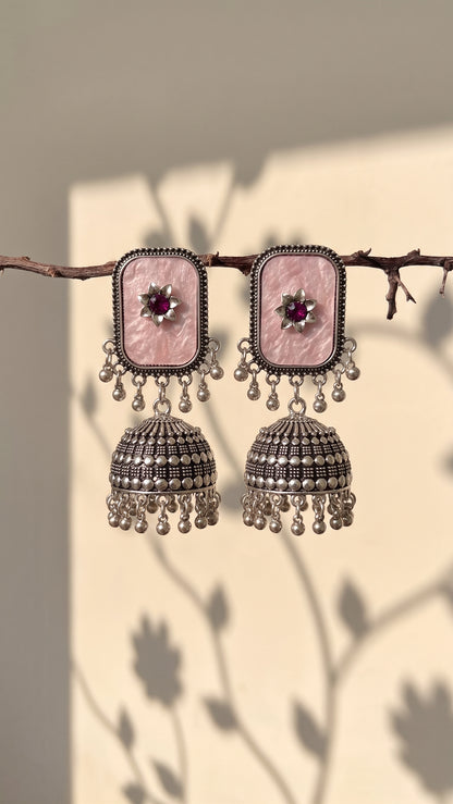 A pair of traditional Indian jhumka earrings with rectangular pink stone studs and silver-toned bell-shaped dangling elements, hanging from a thin branch against a warm beige wall with soft leaf shadows.