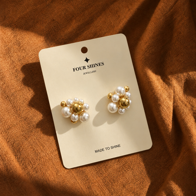A pair of gold and pearl stud earrings displayed on a cream card labeled FOUR SHINES JEWELLERY, placed on a textured rust-brown fabric background.