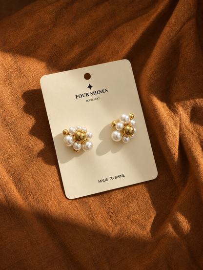 A pair of gold and pearl stud earrings displayed on a cream card labeled FOUR SHINES JEWELLERY, placed on a textured rust-brown fabric background.