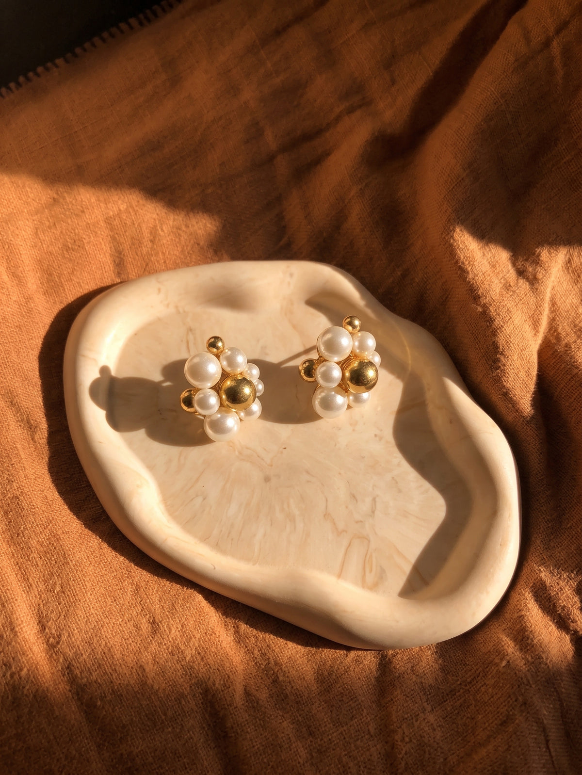 A pair of gold and white pearl stud earrings displayed on a small organic-shaped cream tray, set against a draped brown fabric background with warm sunlight and shadows.