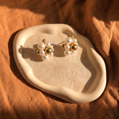 A pair of gold and white pearl stud earrings displayed on a small organic-shaped cream tray, set against a draped brown fabric background with warm sunlight and shadows.