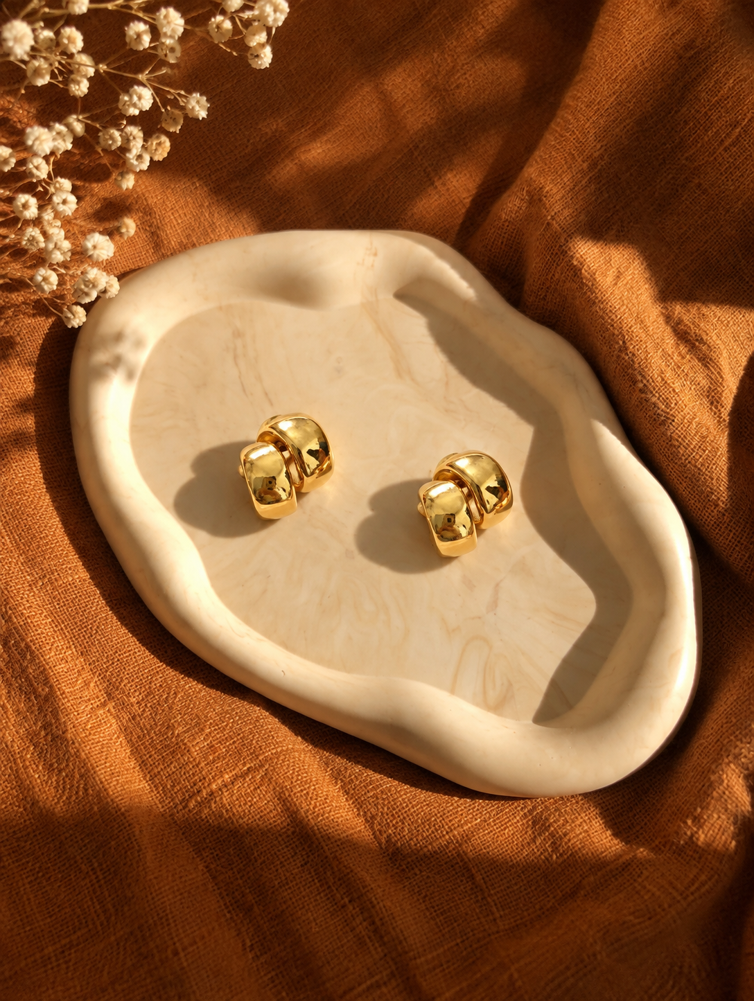 A pair of shiny gold stud earrings displayed on an organic-shaped cream stone tray, set against a warm brown linen fabric background with dried flowers in the corner.