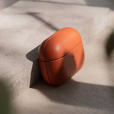 An orange leather wireless earbud charging case resting on a light concrete surface with natural shadows.
